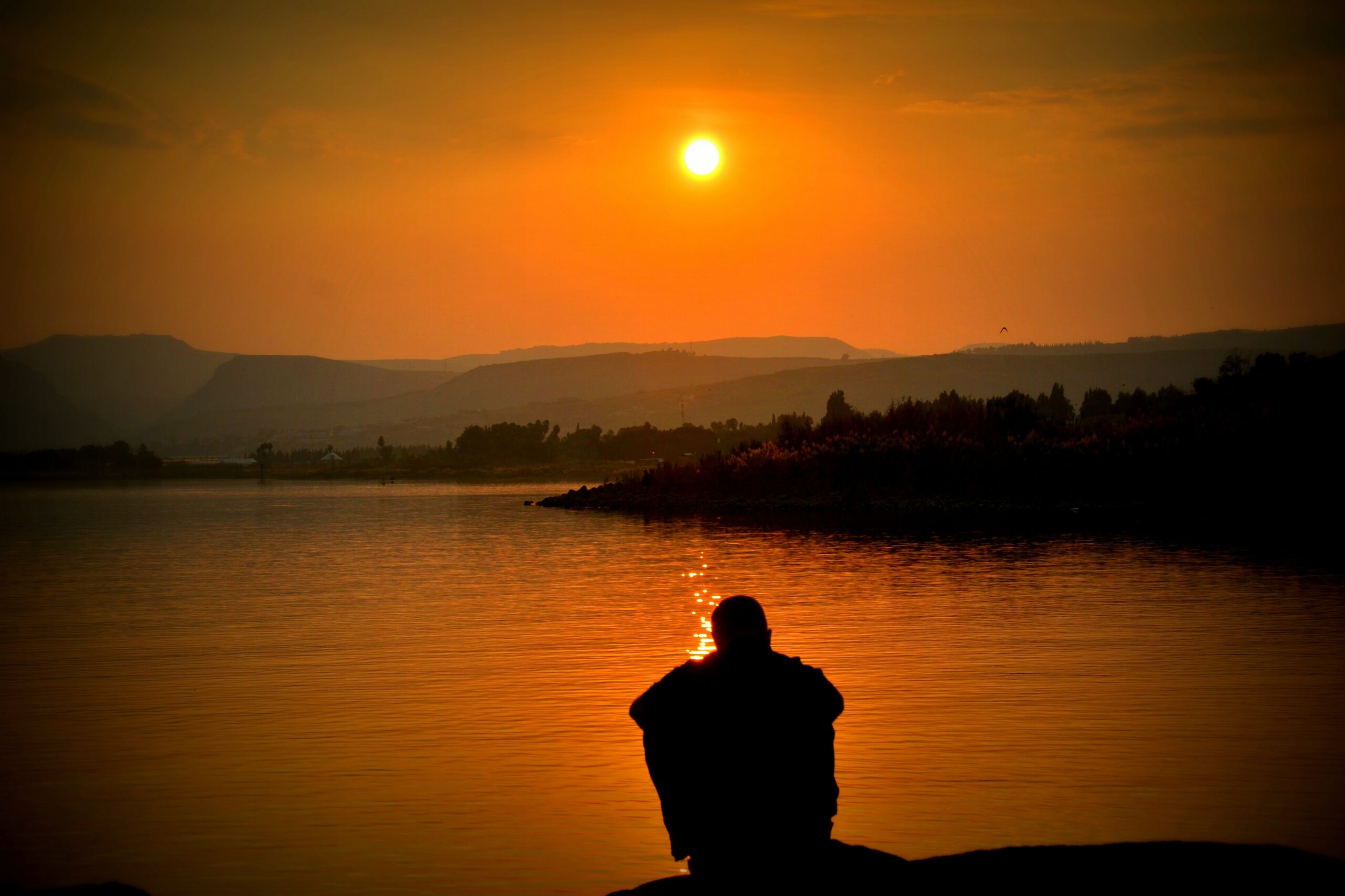 Människa som sitter vid sjön och ser ner mot en orange solnedgång